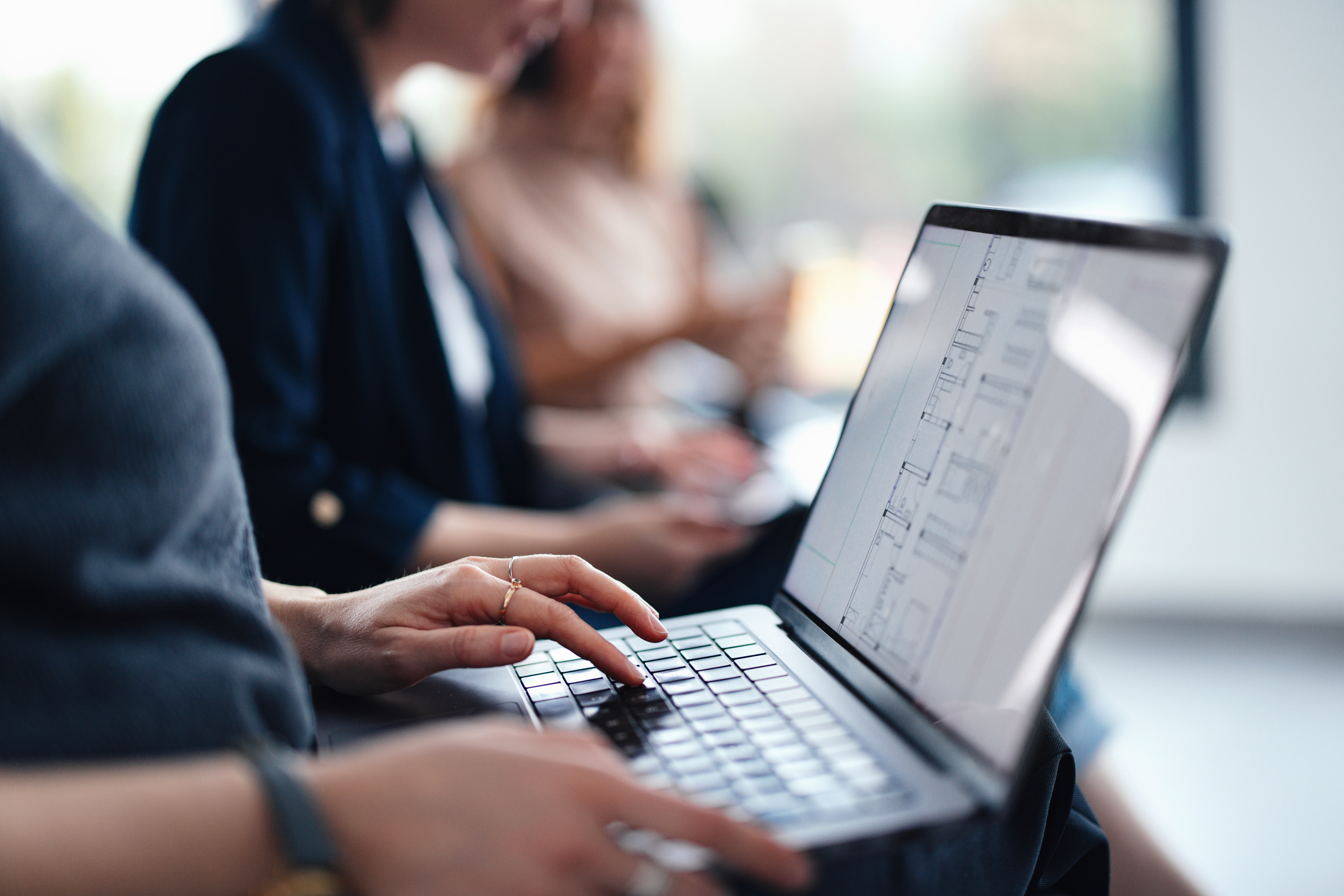 Hands of a person typing on a laptop, in a professional setting, with individuals in the background participating in a collaborative, work-related task, emphasizing productivity and teamwork.